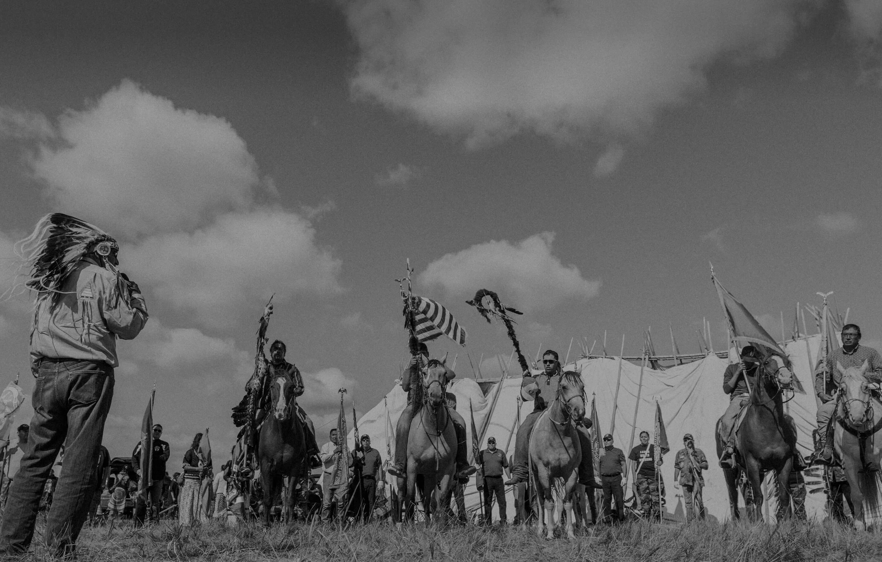 A low angle black and white photograph of Indigenous people on horseback with a large tipi and more people standing in the background