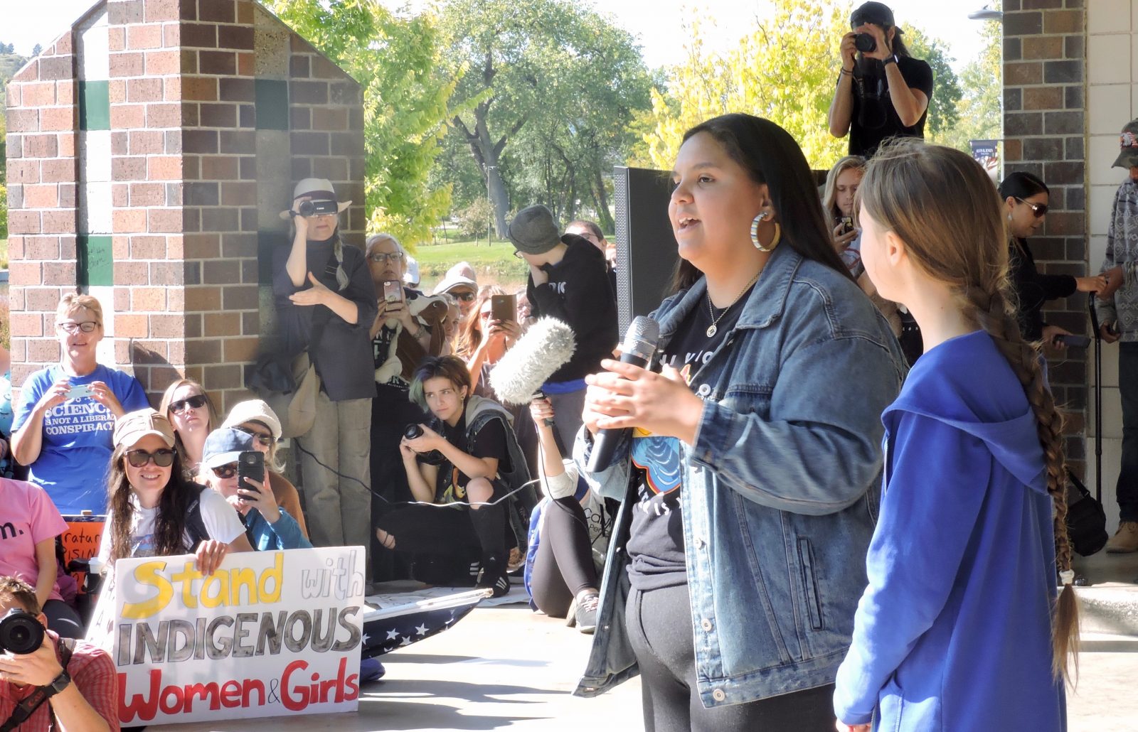 A photograph of young activists Tokata Iron Eyes and Greta Thunberg speaking to a crowd while reporters take photos