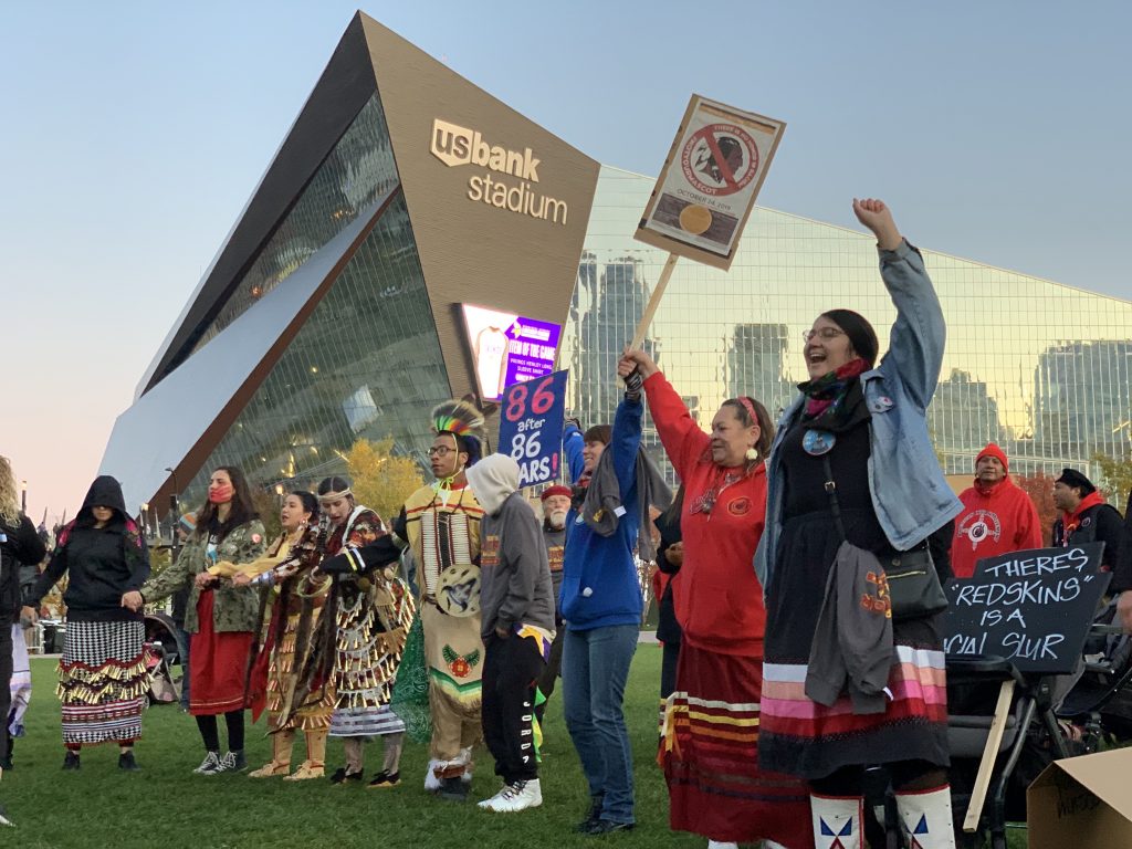 Indigenous people hold protest signs against racist sports mascots in front of US Bank Stadium